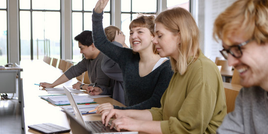 Five students sitting in the seminar room at the table. A female student raises her hand.