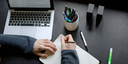 A student working at a table with a laptop, pencils and a notebook.