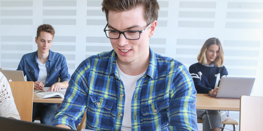 A student is typing on a laptop, two others are sitting behind him.